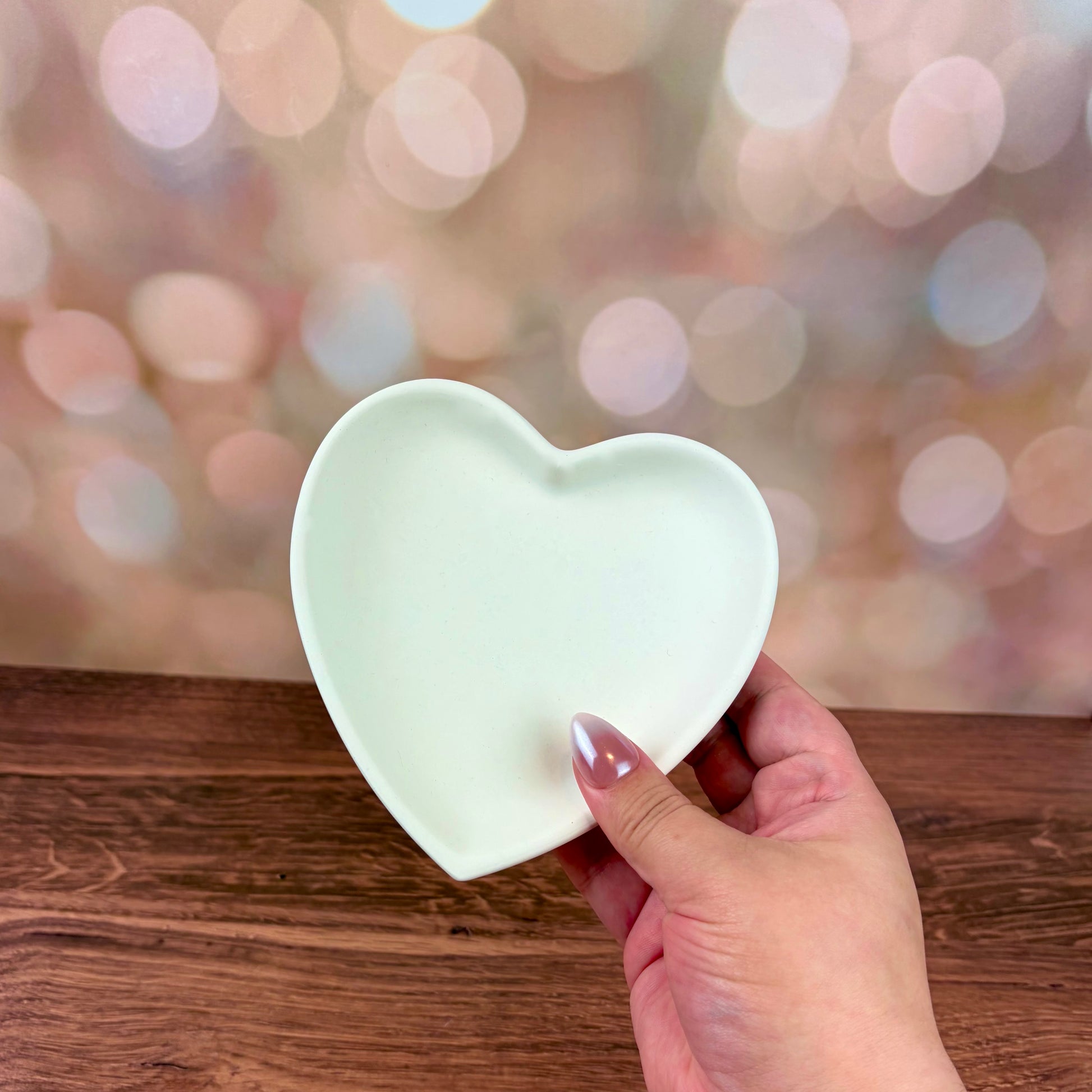 Heart-shaped trinket tray made from white jesmonite held by a hand on a wooden surface with a blurred pink bokeh background.