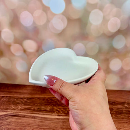 Heart-shaped trinket tray made from white jesmonite held by a hand on a wooden surface with a blurred pink bokeh background.