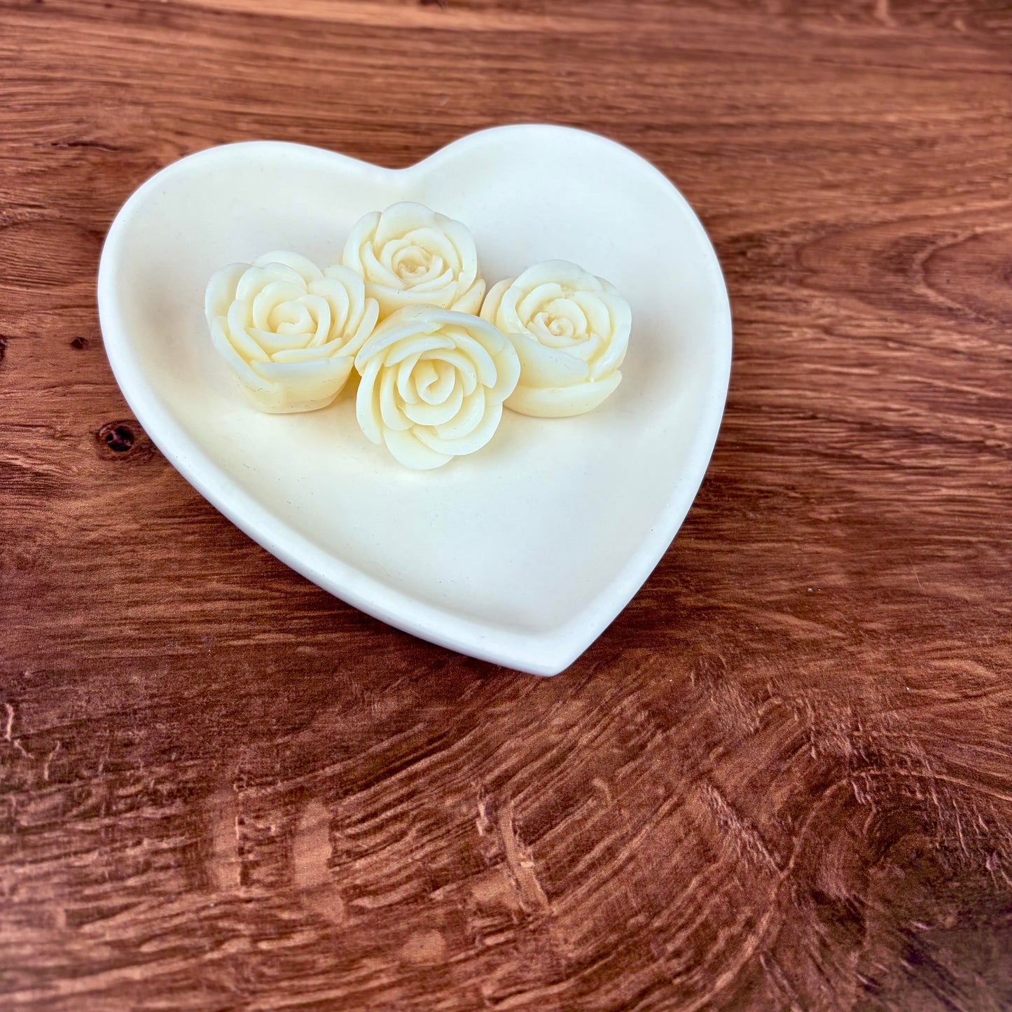 4 white rose wax melts sat in a heart-shaped trinket tray made from white jesmonite sat on a wooden surface with a blurred pink bokeh background.