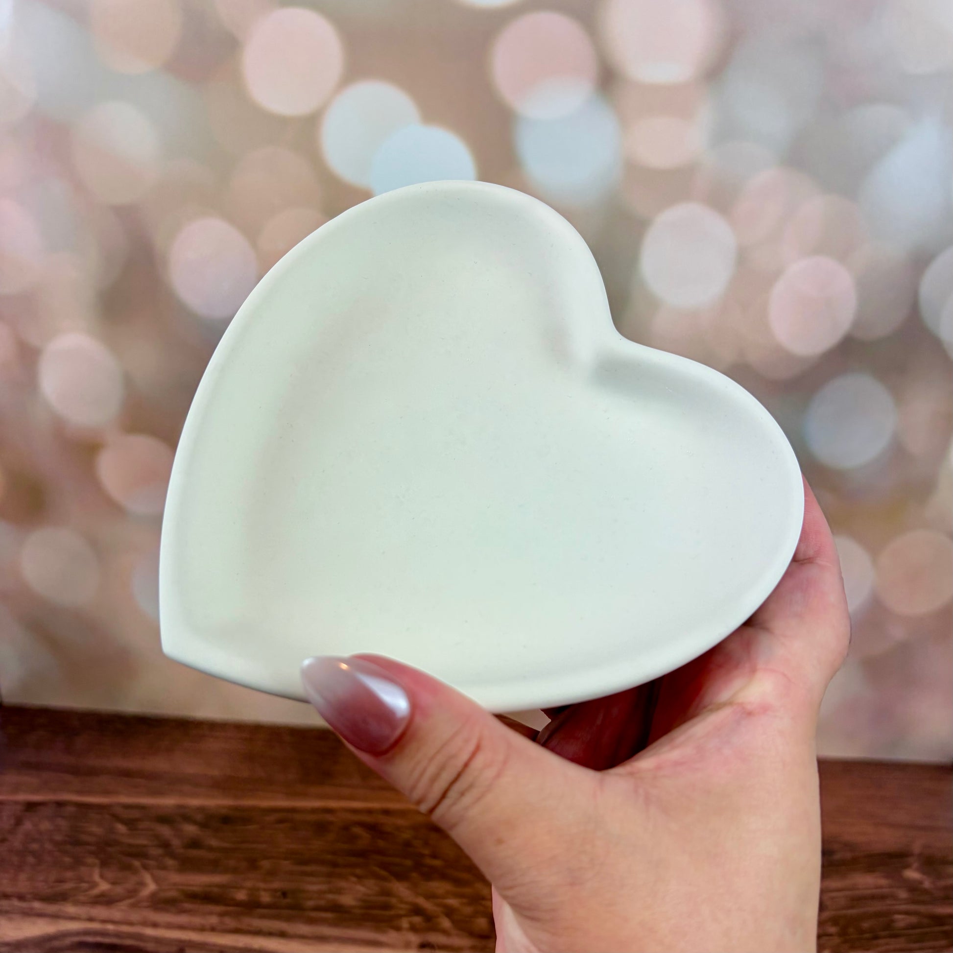 Heart-shaped trinket tray made from white jesmonite held by a hand on a wooden surface with a blurred pink bokeh background.