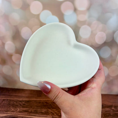 Heart-shaped trinket tray made from white jesmonite held by a hand on a wooden surface with a blurred pink bokeh background.