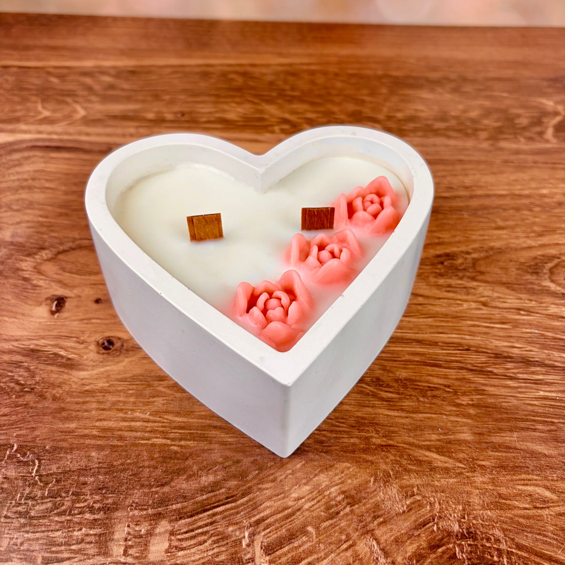 Heart-shaped double wicked candle with 3 pink floral sculpted wax shaped sat inside the white wax candle, accompanied by 2 wooden wicks. This romantic candle sits on a wooden surface.