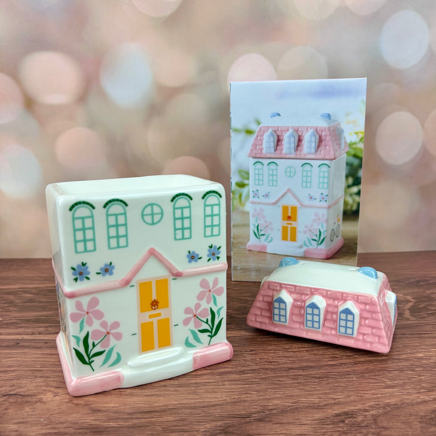 White house-shaped oil burner and wax warmer, featuring a yellow door, floral motifs in a 3 storey regency style. Burner sits on a wooden surface with box behind and pink tiled roof lid sitting in front, against a blurred background