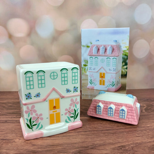 White house-shaped oil burner and wax warmer, featuring a yellow door, floral motifs in a 3 storey regency style. Burner sits on a wooden surface with box behind and pink tiled roof lid sitting in front, against a blurred background