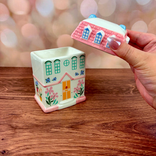 White house-shaped oil burner and wax warmer, featuring a yellow door, floral motifs in a 3 storey regency style. Burner sits on a wooden surface with pink tiled roof lid in hand against a blurred background.