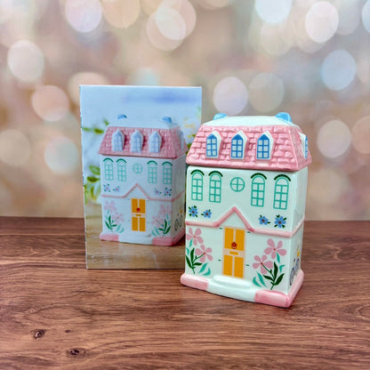 White house-shaped oil burner and wax warmer, featuring pink tiled roof, a yellow door, floral motifs in a 3 storey regency style. Burner and matching box sits on a wooden surface against a blurred background.