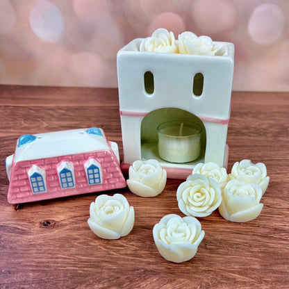 White house-shaped oil burner and wax warmer, featuring pink tiled roof, a yellow door, floral motifs in a 3 storey regency style. Burner and rose wax melts sit on a wooden surface against a blurred background.