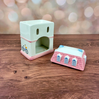 Back of white house-shaped oil burner and wax warmer, featuring a domed opening for the tealight and two air holes, accompanied by the pink tiled roof, both sitting on a wooden surface against a blurred background.