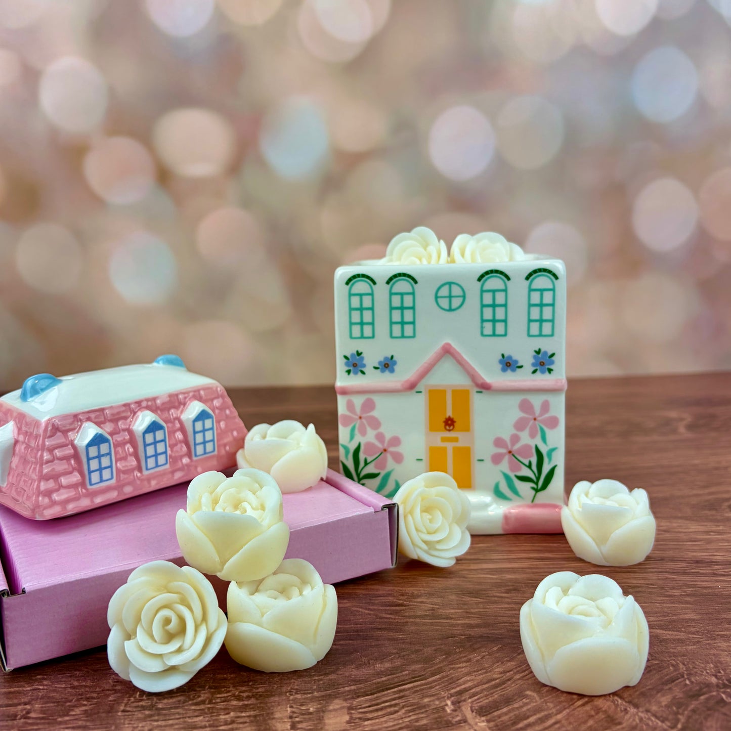 White house-shaped oil burner and wax warmer, featuring pink tiled roof, a yellow door, floral motifs in a 3 storey regency style. Burner, sits next to pink box with roof and rose wax melts on a wooden surface against a blurred background.