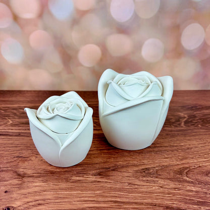 White rose-shaped candles, in two sizes with sculpted petal rose lids, both sat on a wooden surface with a blurred pink bokeh background
