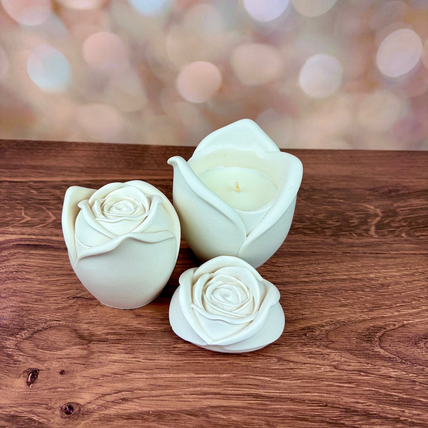 White rose-shaped candles, in two sizes with sculpted petal rose lids, the larger lid is placed in front of the large candle. Both candles are sat on a wooden surface with a blurred pink bokeh background
