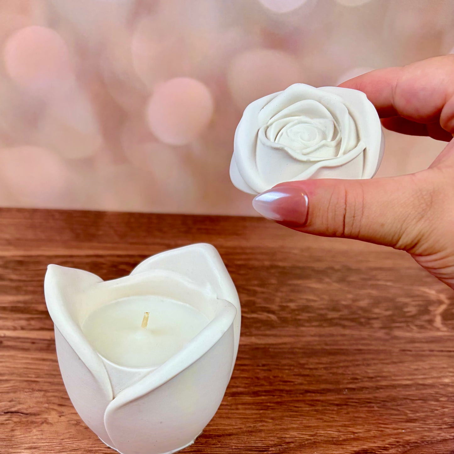 White rose-shaped candle, with sculpted petal rose lid held above the candle, sat on a wooden surface with a blurred pink bokeh background.