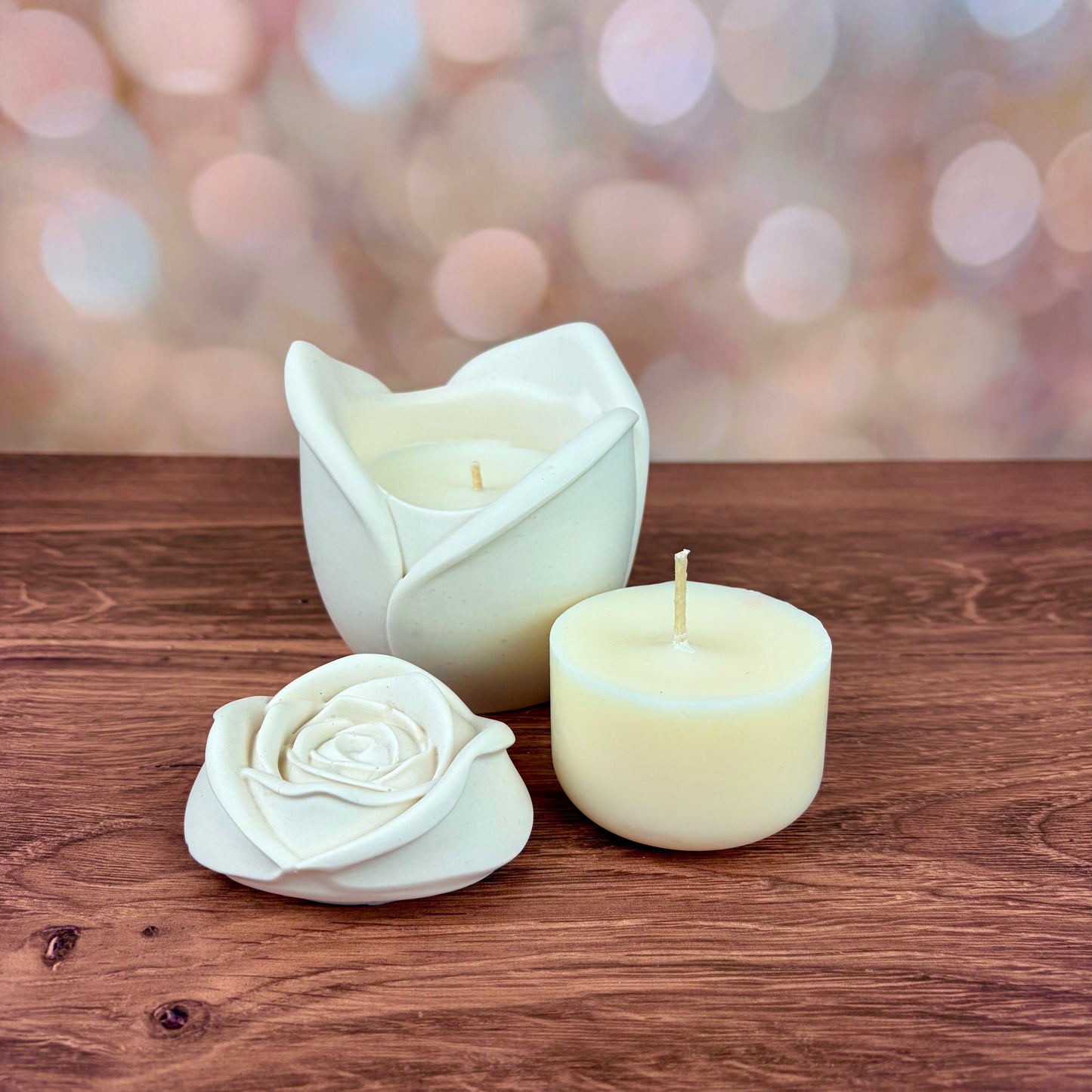 White rose-shaped candles, with sculpted petal rose lid and candle refill placed in front of the candle.  Candles sat on a wooden surface with a blurred pink bokeh background.