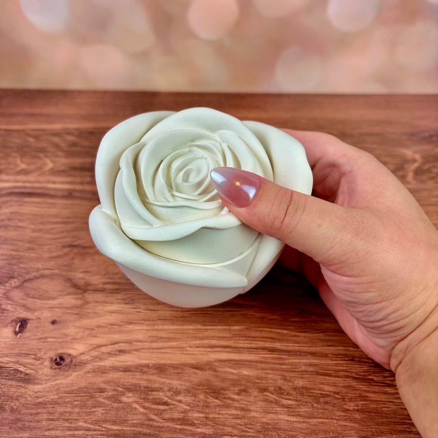 White rose-shaped candle, in large size with sculpted petal rose lid, held over a wooden surface with a blurred pink bokeh background