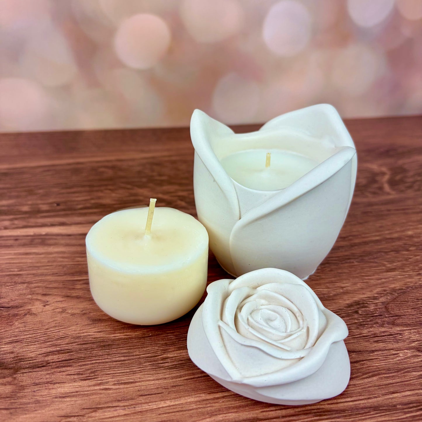 White rose-shaped candles, with sculpted petal rose lid and candle refill placed in front of the candle.  Candles sat on a wooden surface with a blurred pink bokeh background.