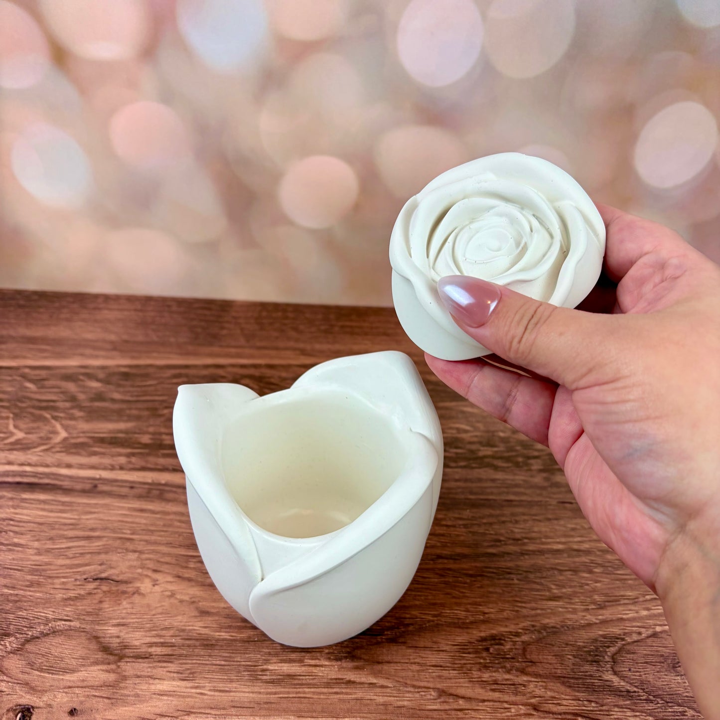 White rose-shaped candle vessel, with sculpted petal rose lid held above the candle, sat on a wooden surface with a blurred pink bokeh background.