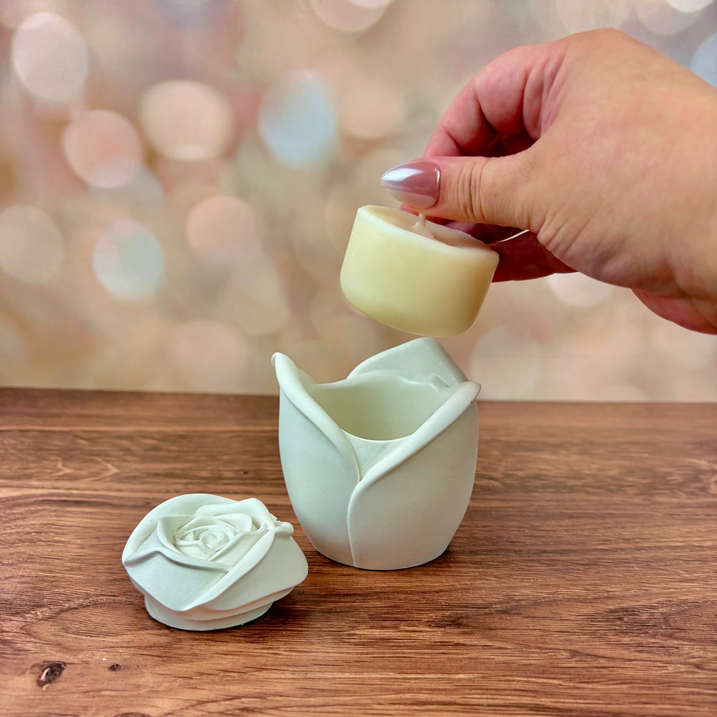White rose-shaped candle, with sculpted petal rose lid in front of candle vessel, and refill held above the candle vessel. These are sat on a wooden surface with a blurred pink bokeh background.