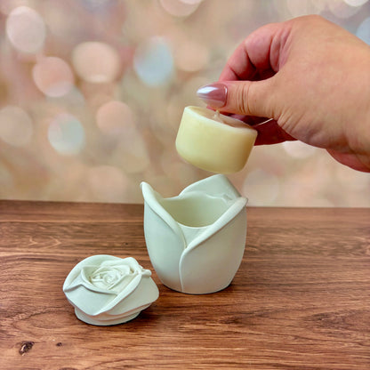 White rose-shaped candle, with sculpted petal rose lid in front of candle vessel, and refill held above the candle vessel. These are sat on a wooden surface with a blurred pink bokeh background.