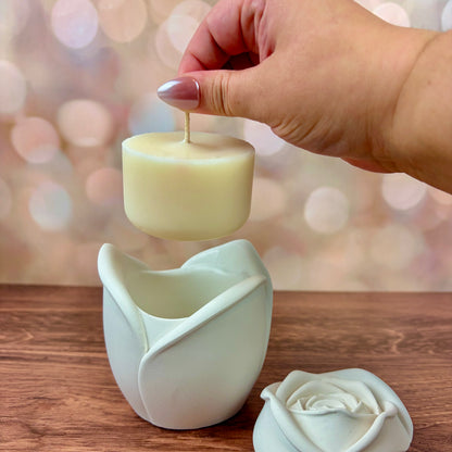 White rose-shaped candle, with sculpted petal rose lid in front, and refill held above the candle, sat on a wooden surface with a blurred pink bokeh background.