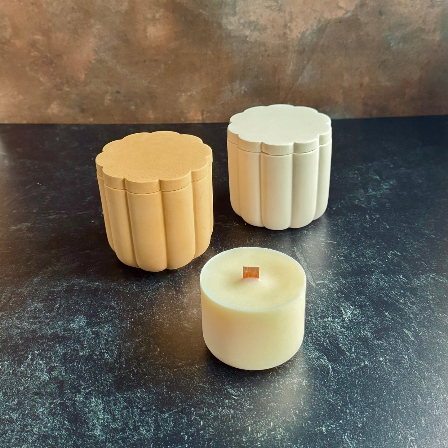 A white and peach pair of scalloped candle vessels with lids, and a wooden wicked candle refill, all sat on a dark surface with a neutral background.