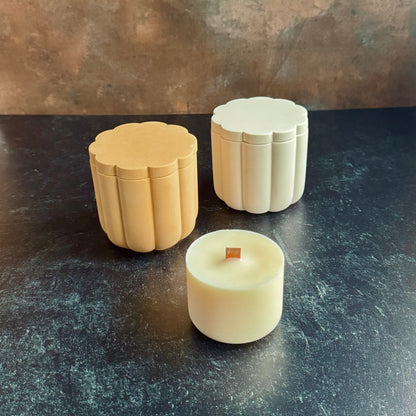 A white and peach pair of scalloped candle vessels with lids, and a wooden wicked candle refill, all sat on a dark surface with a neutral background.
