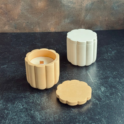 Two scalloped candle vessels with lids, and wooden wicked candle refill. A white scalloped vessel with lid on, and one peach with a lid placed in front showcase the wooden wick. Both sat on a dark surface with a neutral background.