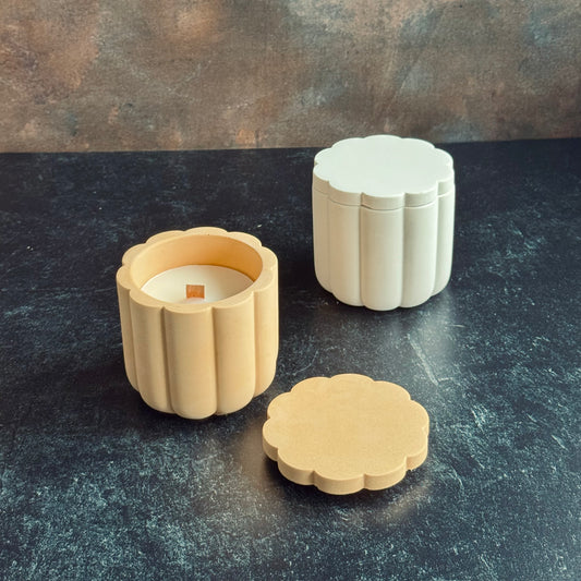 Two scalloped candle vessels with lids, and wooden wicked candle refill. A white scalloped vessel with lid on, and one peach with a lid placed in front showcase the wooden wick. Both sat on a dark surface with a neutral background.