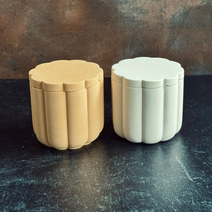 A white and peach pair of scalloped candle vessels with lids, sat on a dark surface with a neutral background.