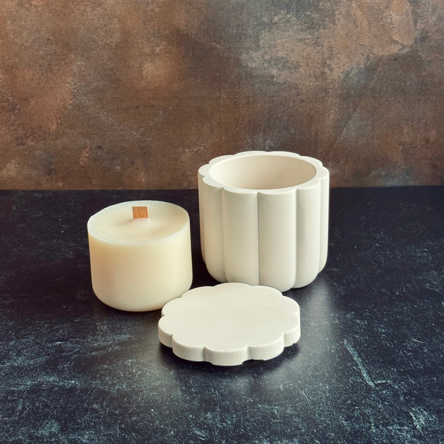 A white scalloped candle vessel with lid sat in front next to a wooden wicked candle refill, all sat on a dark surface with a neutral background.