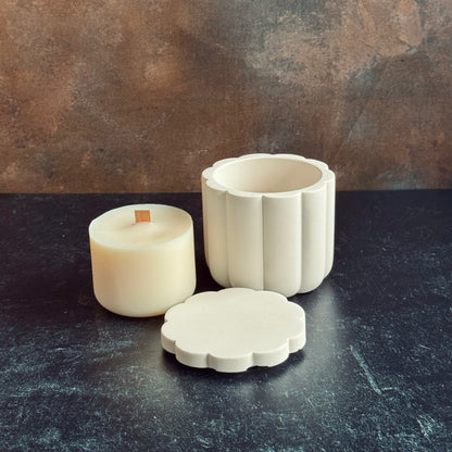A white scalloped candle vessel with lid sat in front next to a wooden wicked candle refill, all sat on a dark surface with a neutral background.