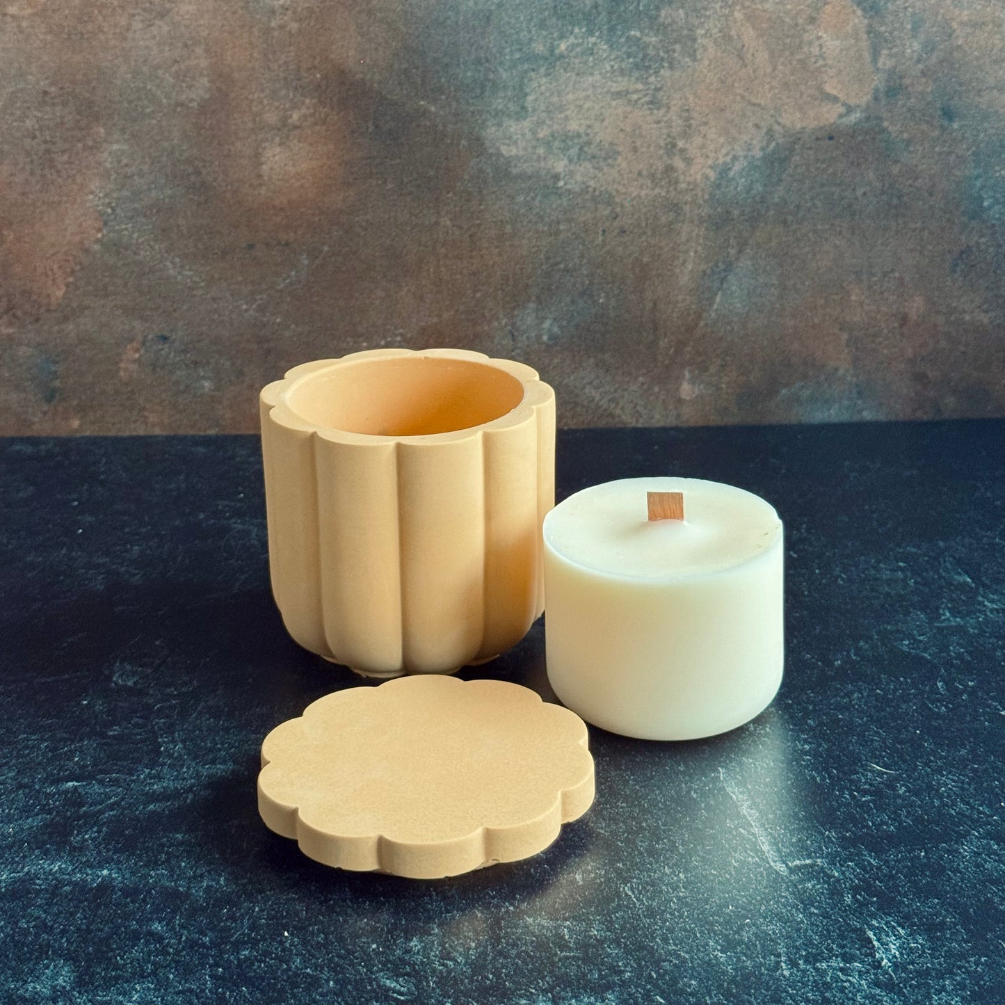 A peach scalloped candle vessel with lid sat in front next to a wooden wicked candle refill, all sat on a dark surface with a neutral background.