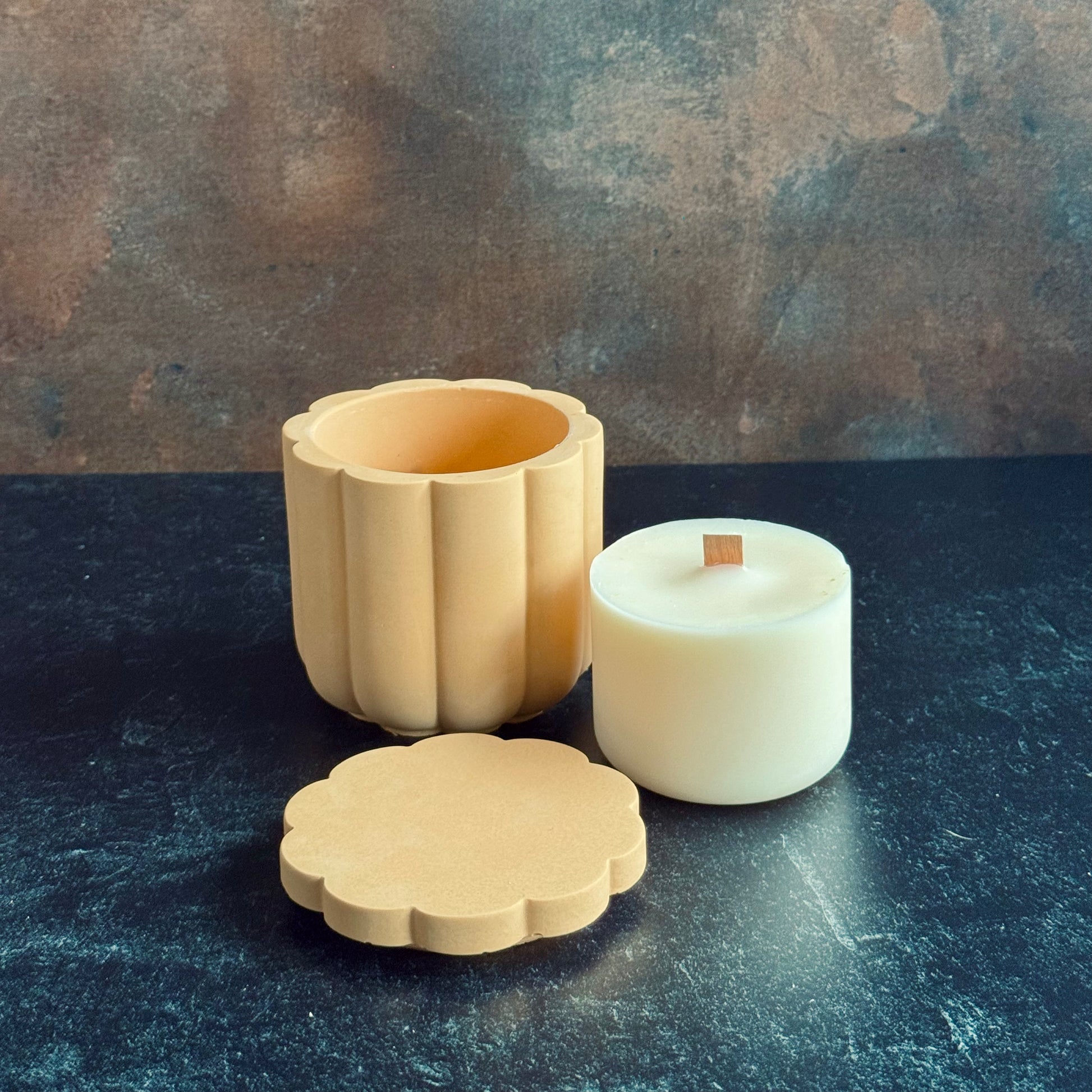 A peach scalloped candle vessel with lid sat in front next to a wooden wicked candle refill, all sat on a dark surface with a neutral background.